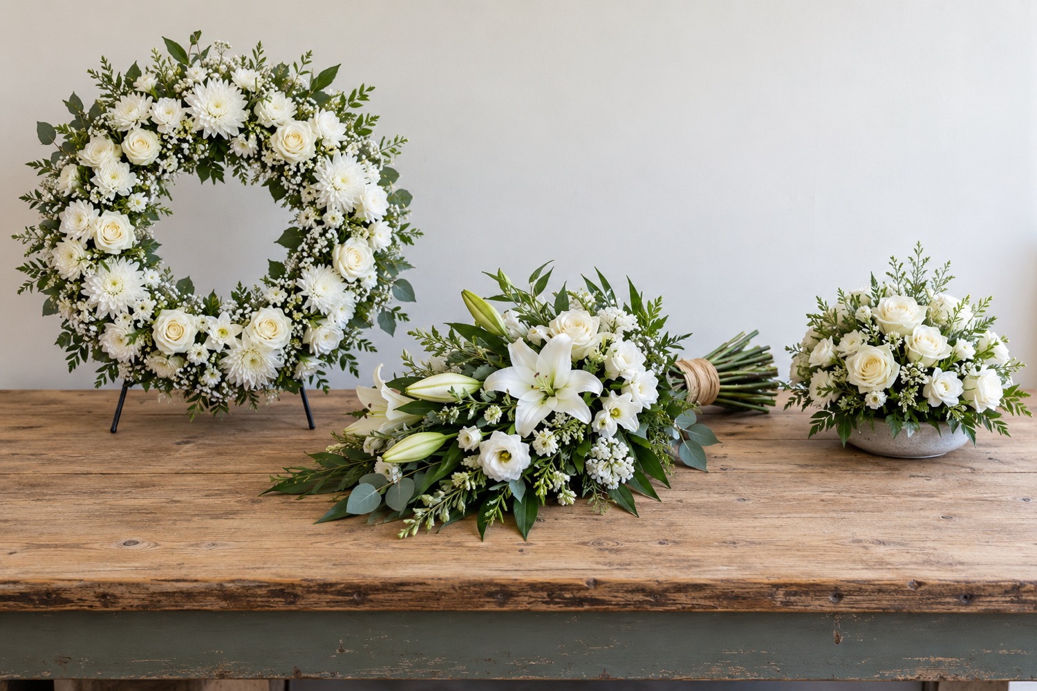 White floral wreath, flat tied sheaf and low posy displayed on a florist workbench.