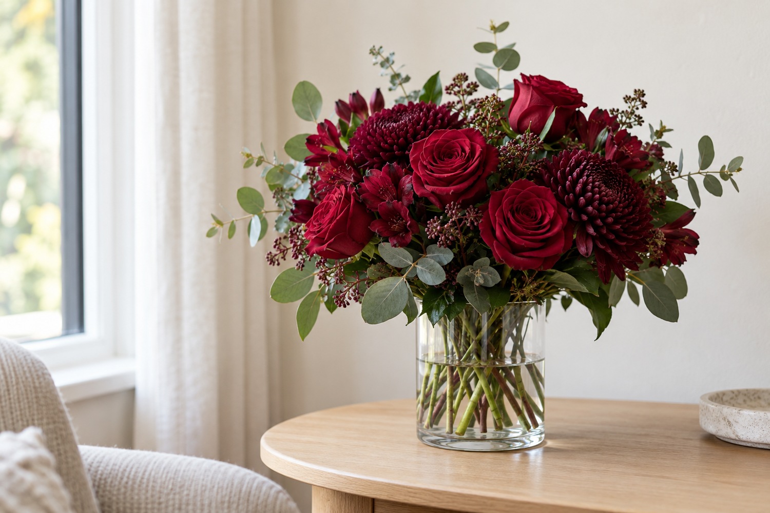 Deep burgundy sympathy flowers in a clear vase beside a window.